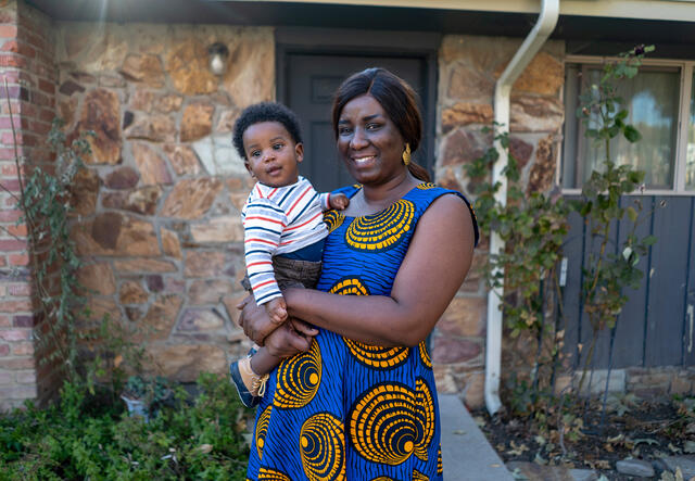 A woman in a blue and yellow dress holds her one year old son and smiles while standing in front of a house