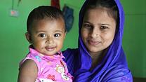 Lovely Akter, an IRC midwife, poses with her nine-month-old daughter. Lovely is wearing a blue headscarf and her daughter is wearing a pink shirt. Both are smiling.